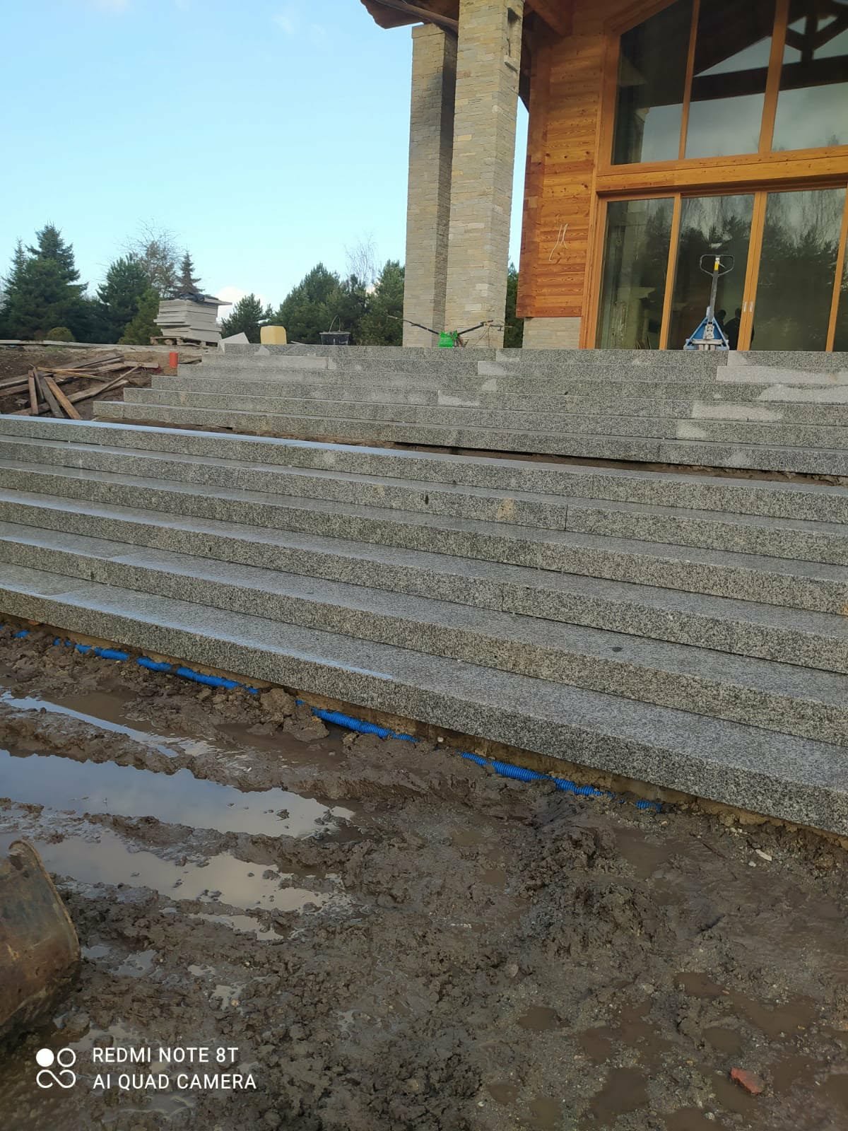 Granitowe schody wejściowe do domu w trakcie budowy, widoczne niebieskie rury drenażowe i błoto. Częściowo ułożona kostka granitowa na schodach.