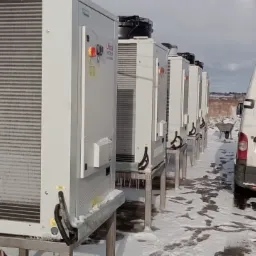 Rząd białych jednostek zewnętrznych na metalowych stelażach, prawdopodobnie urządzeń grzewczych, ustawionych na zewnątrz w zimowych warunkach, obok zaparkowanego białego samochodu dostawczego...