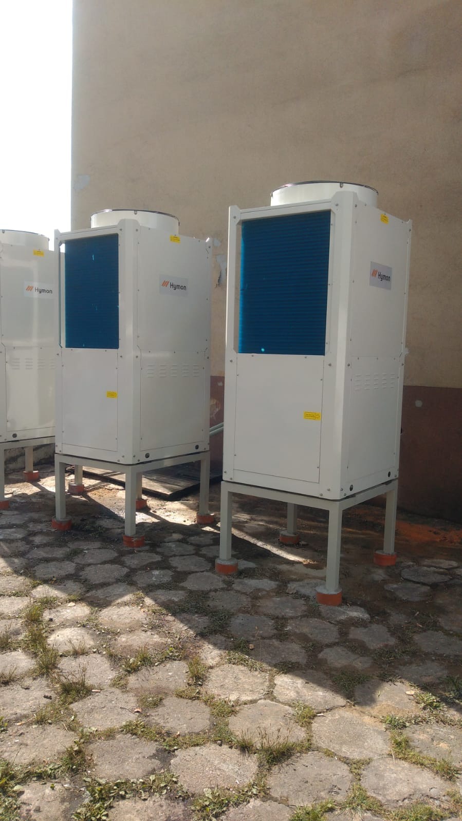 Trzy identyczne, białe pompy ciepła marki Hymon, ustawione na zewnątrz budynku, na betonowej nawierzchni z widocznymi chwastami, z pomarańczowymi elementami na podstawie.