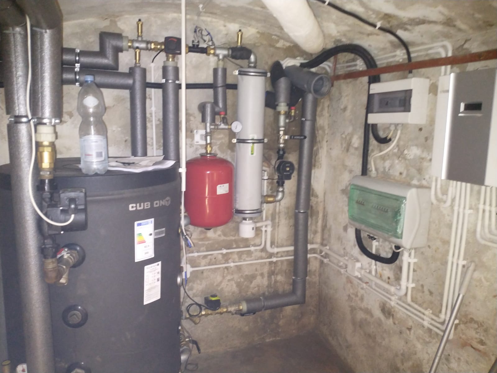 Kotłownia z czarnym zbiornikiem CUB ON, czerwonym naczyniem przeponowym, izolowanymi rurami, białą butelką wody na zbiorniku i skrzynkami elektrycznymi na ścianie z surowego betonu.