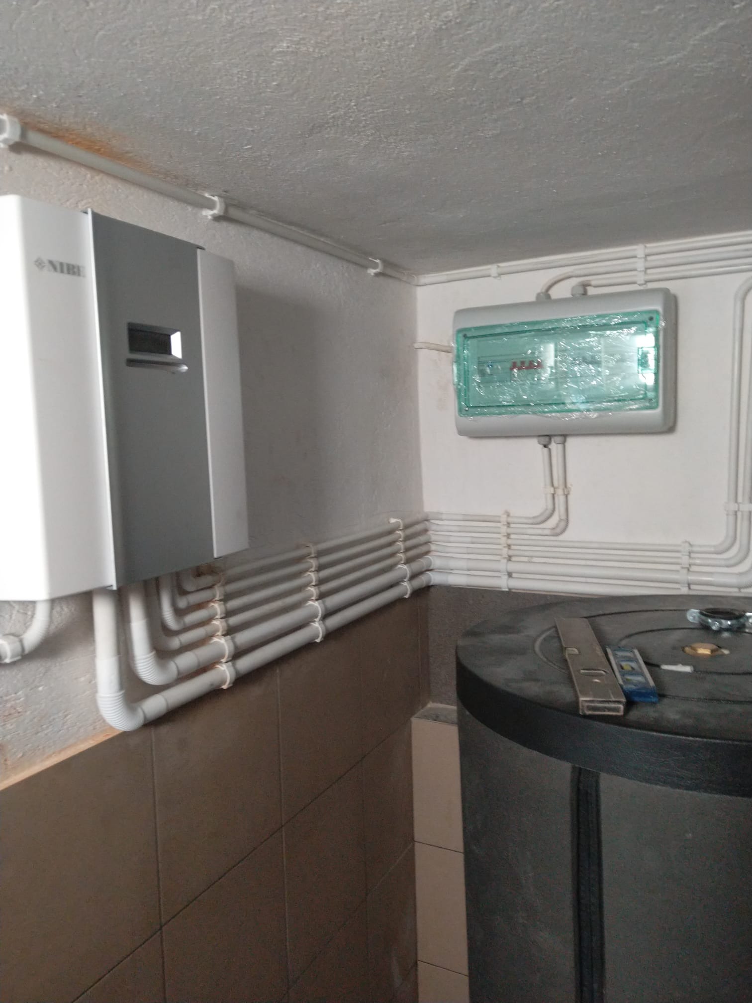 Szara jednostka grzewcza NIBE zamontowana na ścianie, z podłączonymi szarymi rurami, obok skrzynka elektryczna z zieloną pokrywą zabezpieczoną folią, poziomica na zbiorniku akumulacyjnym, brązowe...