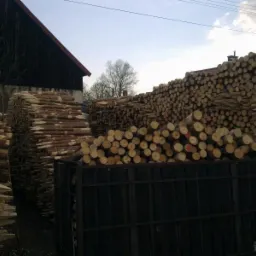 Stosy impregnowanych bali drewnianych na składzie, widoczne końcówki z czerwonymi oznaczeniami, w tle budynek gospodarczy z ciemnego drewna.