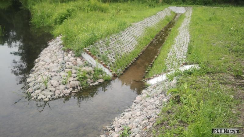 Regulacja koryta rzeki z użyciem gabionów i betonowych elementów. Brzeg umocniony kamieniami w siatce. Widoczne rośliny i woda. Prace inżynieryjne.