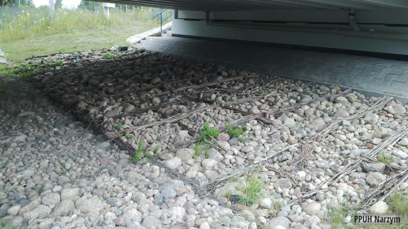 Gabiony z kamienia pod mostem w Iłowo-Osadzie. Konstrukcja z siatki wypełniona okrąglakami, zabezpieczająca skarpę. Widoczne elementy konstrukcji mostu.