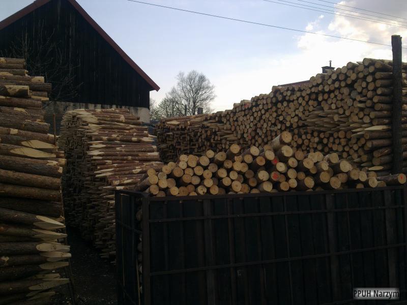 Stosy impregnowanych bali drewnianych na składzie, widoczne końcówki z czerwonymi oznaczeniami, w tle budynek gospodarczy z ciemnego drewna.