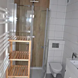 Nowoczesna łazienka z kabiną prysznicową typu walk-in z drewnianym panelem, drewnianym regałem, białą toaletą podwieszaną i umywalką z szafką.