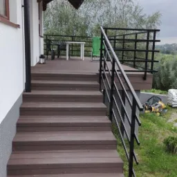 Zadaszony taras z brązowych desek kompozytowych z czarną metalową balustradą i schodami, widok z dołu na meble ogrodowe i zielone otoczenie.