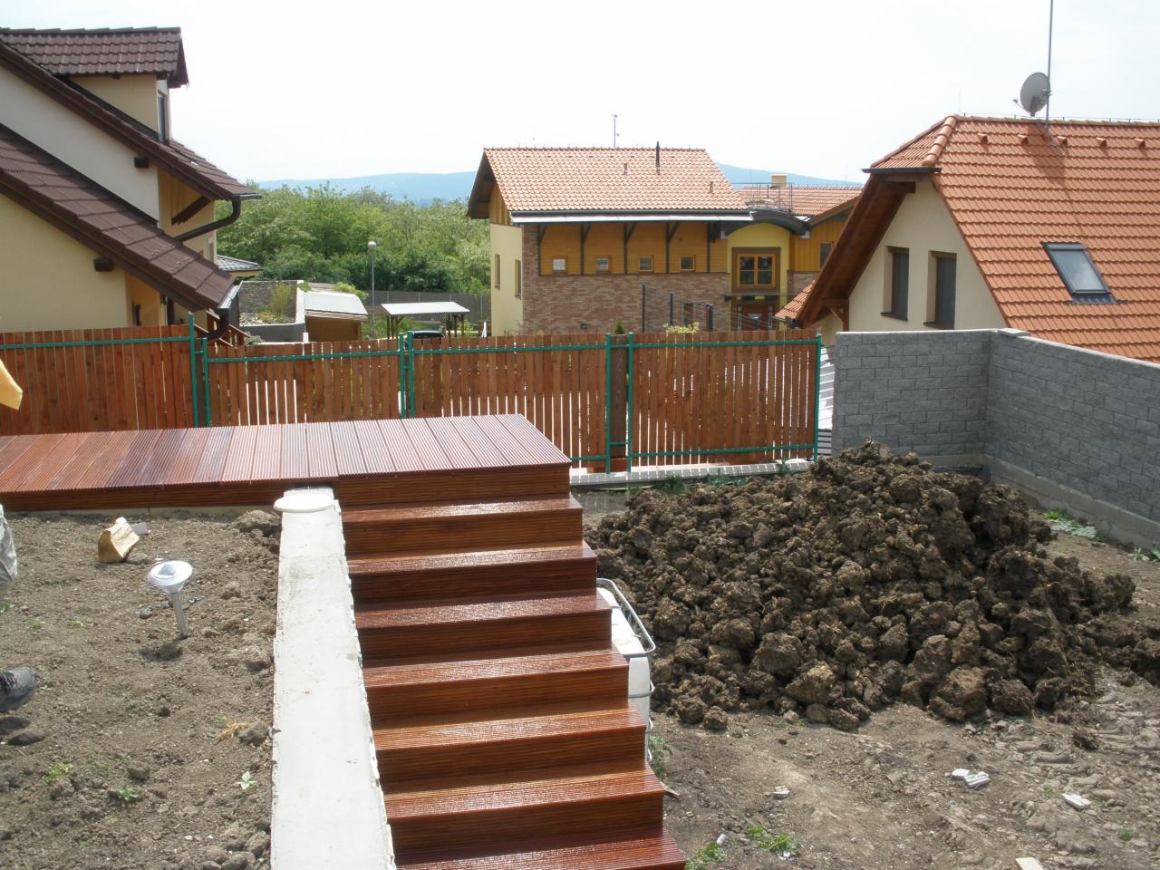 Drewniany taras w trakcie budowy z widocznymi schodami i ogrodzeniem w tle, obok pryzma ziemi.