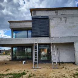 Surowy budynek w trakcie montażu zewnętrznych żaluzji fasadowych w kolorze antracytowym, widoczne drabiny opierające się o ścianę, okna z widocznym odbiciem nieba.