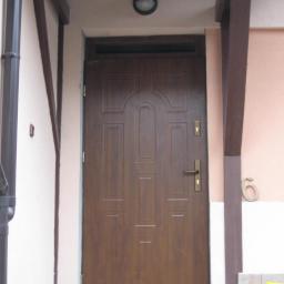 WADEX - Brązowe drzwi wejściowe z dekoracyjnymi panelami, zamontowane w ościeżnicy z drewnianymi belkami, widoczny numer domu '6' i koszyk wiklinowy na podłodze.