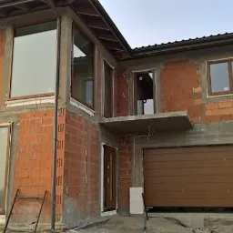 Dom w trakcie budowy z zamontowanymi oknami z brązowymi ramami i brązową bramą garażową, widoczne rusztowania i niezakończone prace murarskie.