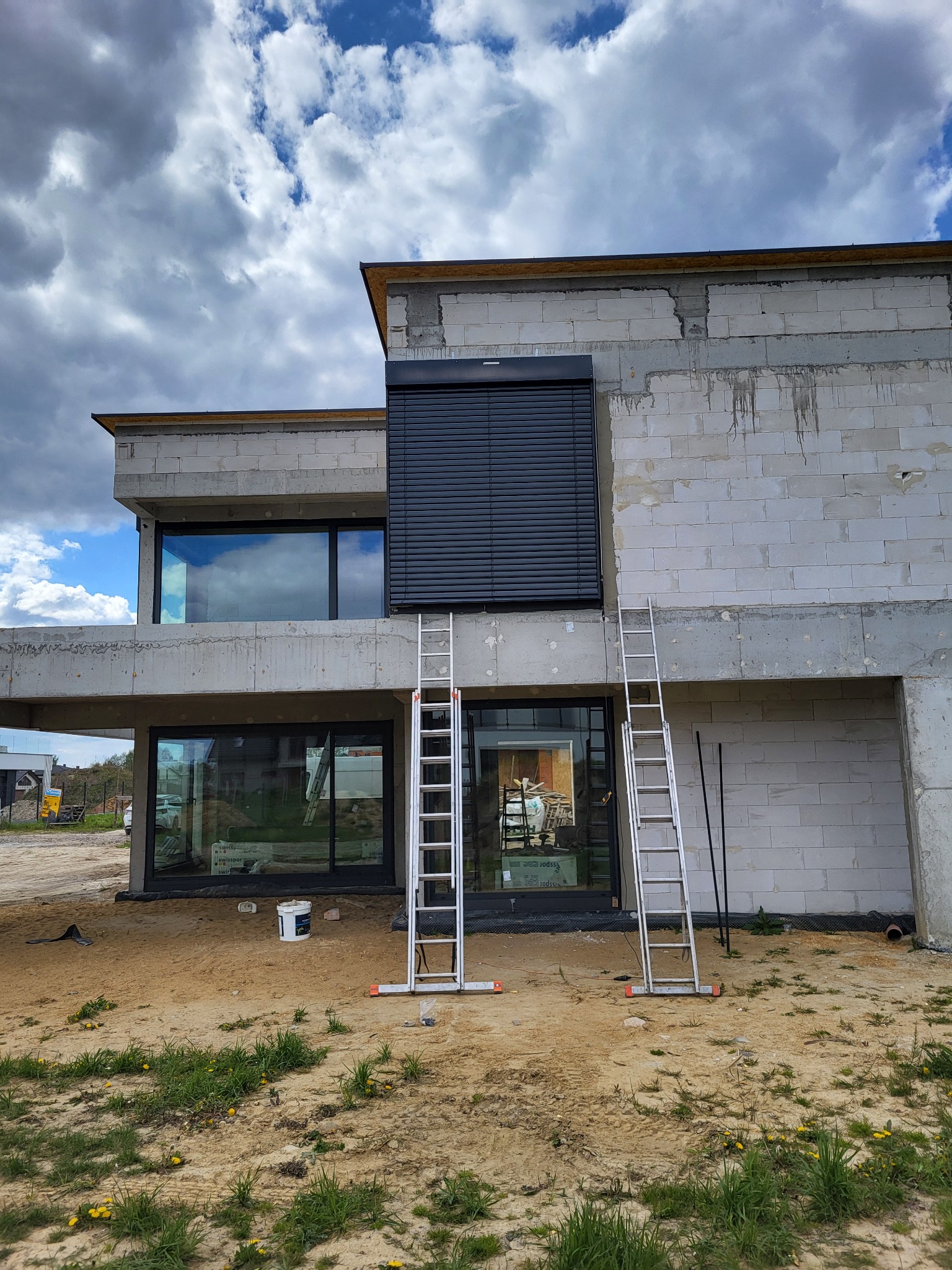 Surowy budynek w trakcie montażu zewnętrznych żaluzji fasadowych w kolorze antracytowym, widoczne drabiny opierające się o ścianę, okna z widocznym odbiciem nieba.