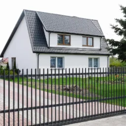 Dom jednorodzinny z ciemnoszarą dachówką karpiówką, białymi ścianami i czarnym ogrodzeniem, widoczny zza furtki, z zadbanym trawnikiem i rabatą kwiatową.