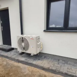 Zewnętrzna jednostka klimatyzacji Viessmann zamontowana na elewacji domu z oknem i ciemnymi drzwiami wejściowymi, widoczna kostka brukowa wokół budynku.