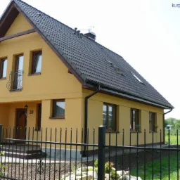 Żółty dom jednorodzinny z ciemnobrązowym dachem, balkonem i oknami, otoczony czarnym metalowym ogrodzeniem i zielenią. Widok z zewnątrz w słoneczny dzień.