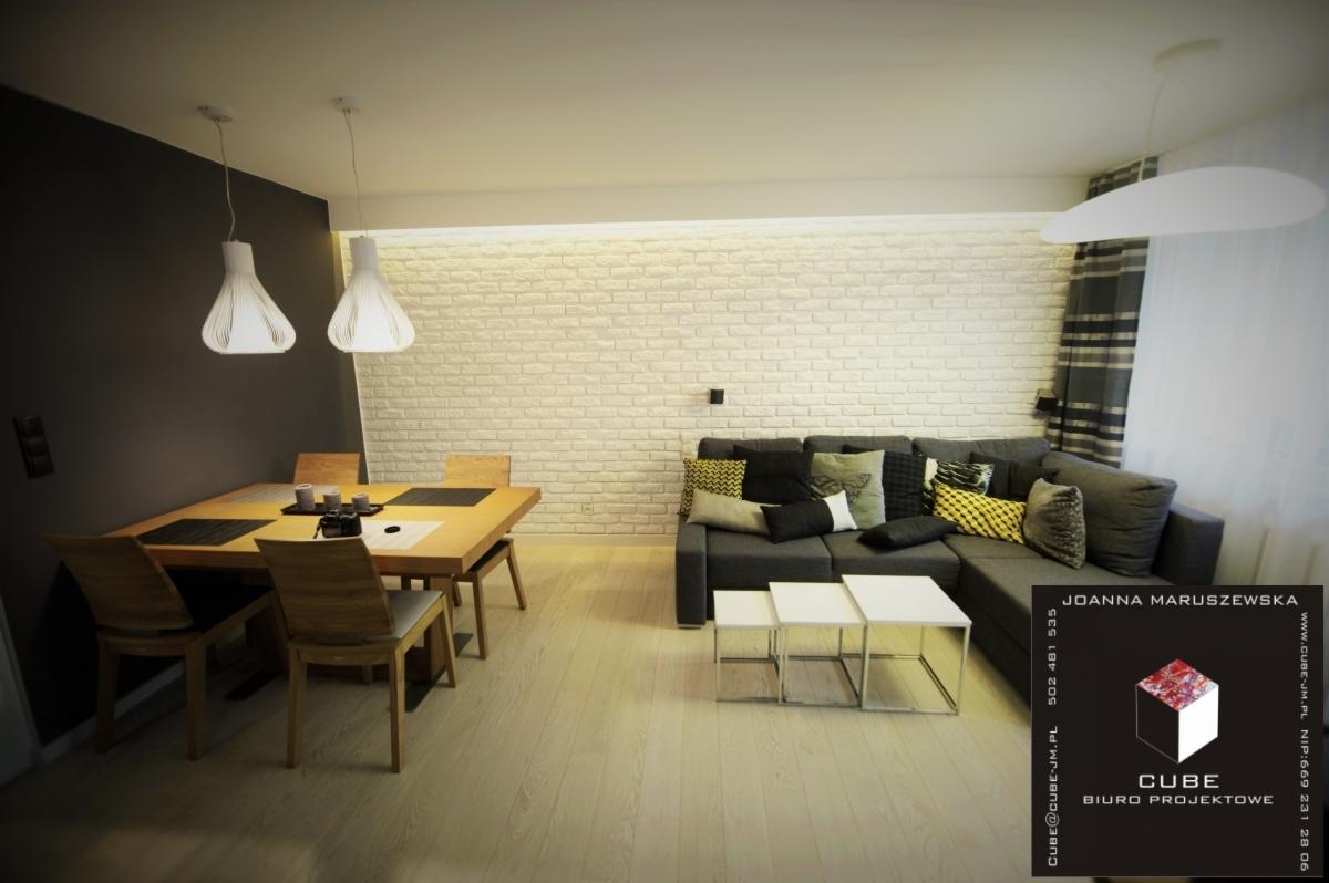 Jasne wnętrze: stół z krzesłami pod lampami, szara sofa z poduszkami na tle białej ściany z cegły. Minimalistyczny design. W rogu logo biura projektowego.