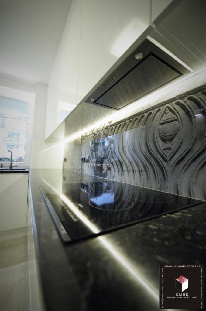 Nowoczesna kuchnia z czarnym blatem i płytą indukcyjną, oświetlona listwą LED pod szafkami. Dekoracyjny panel nad blatem. Widok z boku na okno.