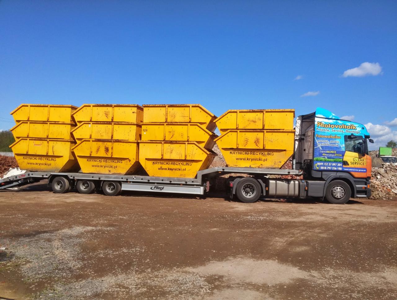 Ciężarówka z żółtymi kontenerami firmy Krynicki Recykling na tle błękitnego nieba. Na kabinie reklama fotowoltaiki. Transport odpadów budowlanych.