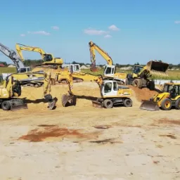 Żółte i szare koparki oraz spycharki różnych marek stojące na piaszczystym placu budowy w słoneczny dzień.