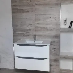 Nowoczesna łazienka z białymi płytkami imitującymi marmur i drewnopodobnymi płytkami. Umywalka z szafką w kolorze białym, toaleta podwieszana i wnęka w ścianie z półkami.