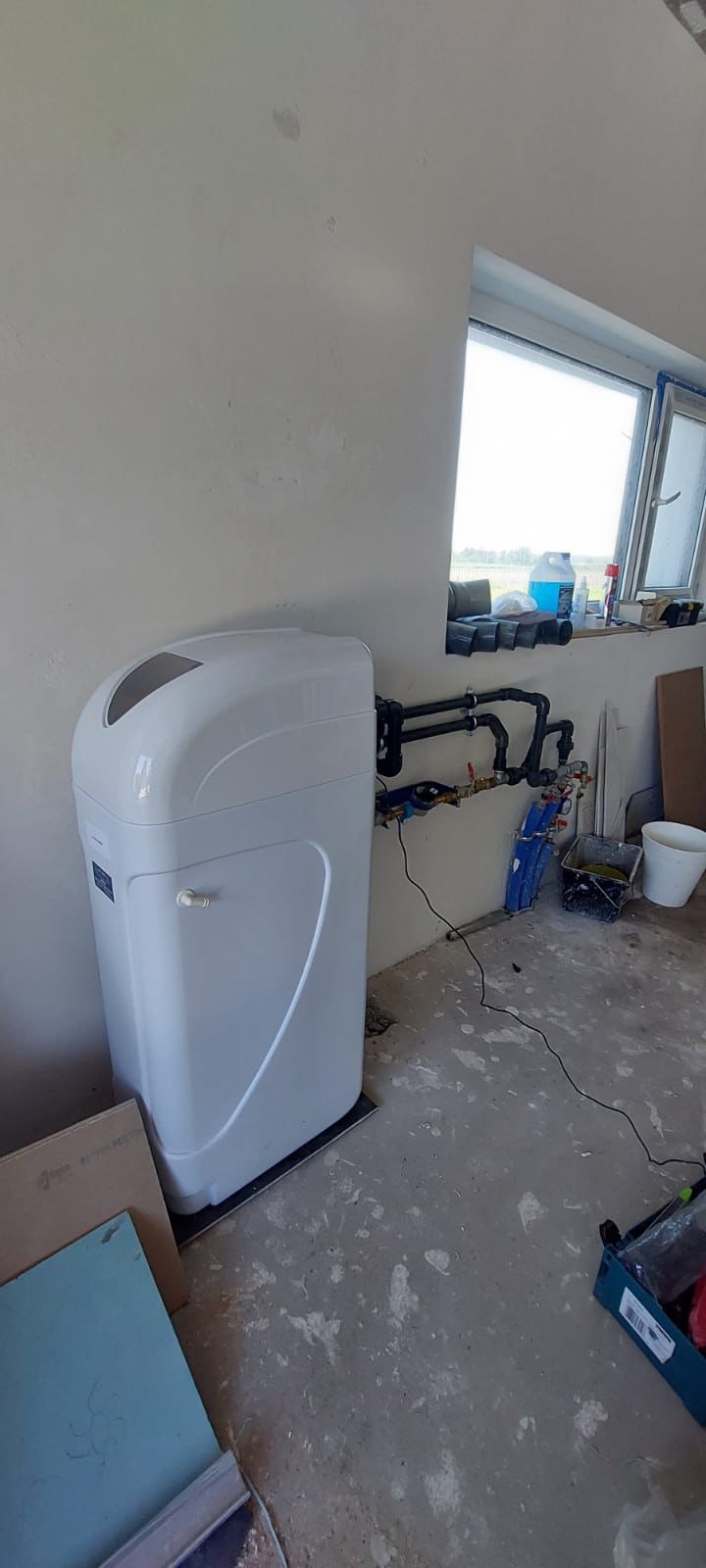 Biały zmiękczacz wody podłączony do instalacji w surowym pomieszczeniu z oknem. Widoczne rury, zawory i narzędzia. Świeża instalacja w trakcie prac wykończeniowych.
