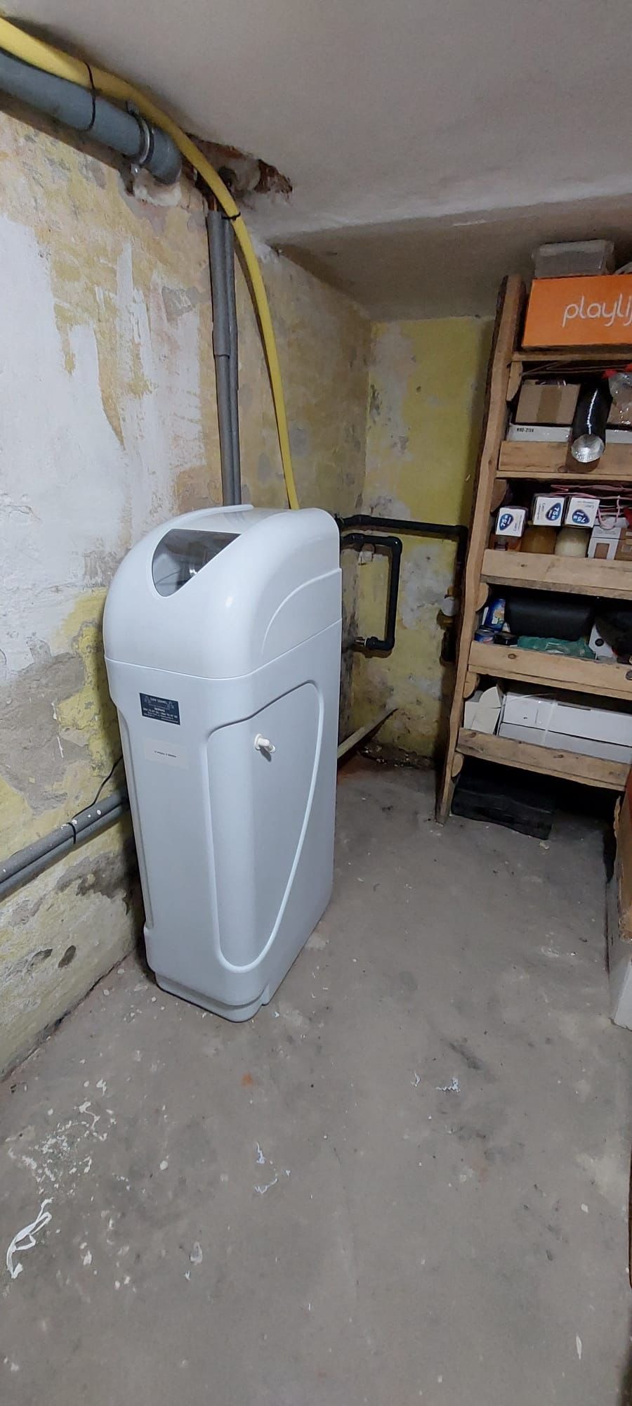Biały zmiękczacz wody w piwnicy z rurami na ścianie i drewnianym regałem. Widoczne przyłącza i instalacja. Ściany z widocznymi śladami użytkowania.