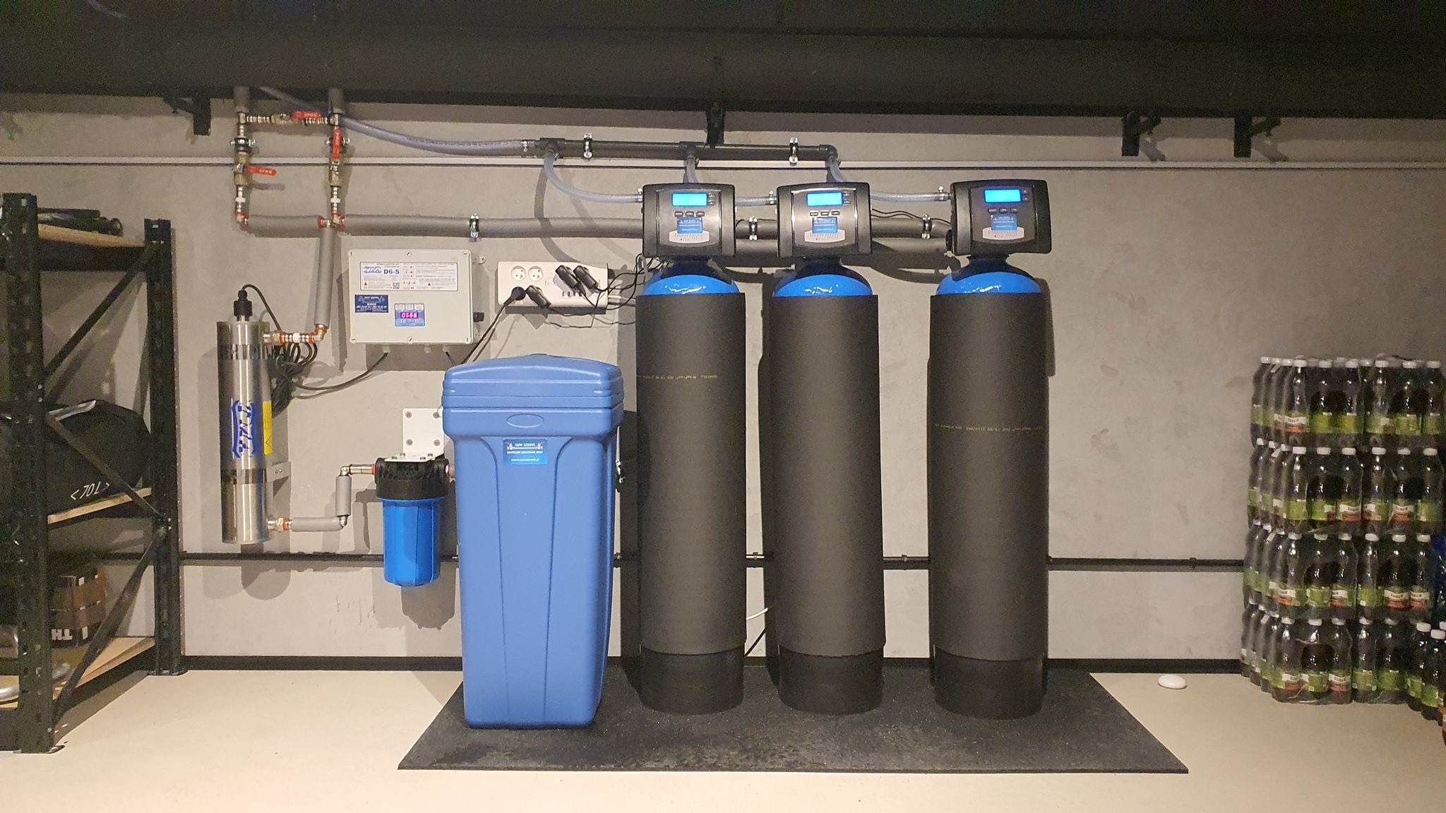 Kompletny system uzdatniania wody w piwnicy: trzy czarne filtry kolumnowe z niebieskimi głowicami, niebieski zmiękczacz i lampa UV na szarej ścianie.