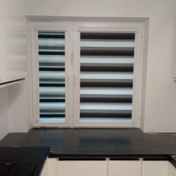 Deco-Light - Dwuskrzydłowe okno z zamontowanymi roletami dzień noc w kolorach szarości, czerni i bieli, widoczne z wnętrza kuchni z białymi szafkami.