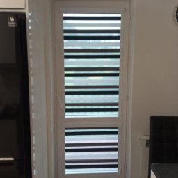 Deco-Light - Okno z zamontowaną roletą dzień noc w kolorze szaro-białym. Widoczne poziome pasy rolety. Ujęcie z bliska, wnętrze mieszkania.