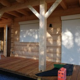 Deco-Light - Białe rolety zewnętrzne na drewnianym domku letniskowym z drewnianą podłogą tarasu, oświetlone popołudniowym słońcem.