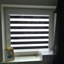 Deco-Light - Okno z zamontowaną roletą zebra w biało-czarne pasy. Na parapecie ramka ze zdjęciem i pudełko w serduszka. Widok od wewnątrz, w tle ściana i zasłona.