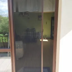 Zamknięta, przesuwna moskitiera plisowana w brązowej ramie, zamontowana w oknie balkonowym. Widok na wnętrze domu przez siatkę moskitiery.