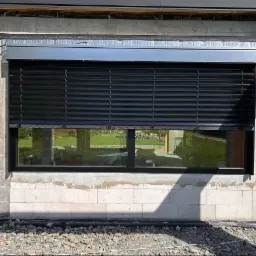 Czarne, poziome osłony okienne na tle surowej ściany z betonu komórkowego, z widoczną instalacją nad oknem i fragmentem krajobrazu w odbiciu szyby.