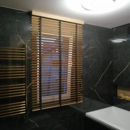 Deco-Light - Beżowe żaluzje drewniane z czarnym sznurkiem w nowoczesnej łazience z czarnym marmurem i złotym grzejnikiem drabinkowym, widok na okno.