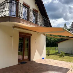 Beżowa markiza tarasowa rozłożona nad brązowymi drzwiami balkonowymi w domu z kutą balustradą. Widok na ogród i sąsiedni budynek w słoneczny dzień.