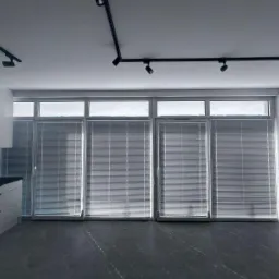 Nowoczesne wnętrze z dużymi oknami przesłoniętymi jasnymi, poziomymi listwami. Minimalistyczny design podkreśla czarna szyna z reflektorami na suficie i błyszcząca zabudowa kuchenna.