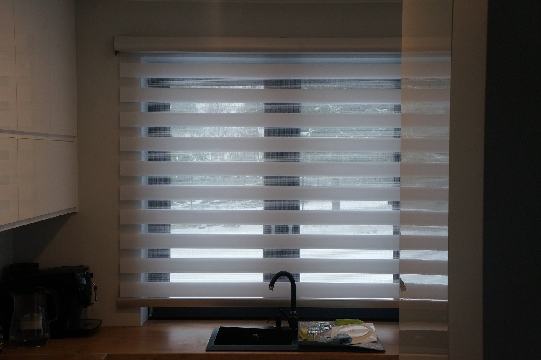 Roleta dzień noc w kuchni, częściowo zasłonięta, z widokiem na zewnątrz. Ujęcie z bliska, ukazujące detale tkaniny i mechanizm rolety. Na blacie zlewozmywak.