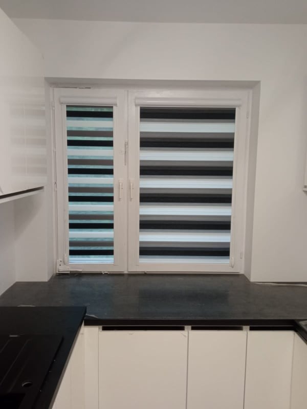 Dwuskrzydłowe okno z zamontowanymi roletami dzień noc w kolorach szarości, czerni i bieli, widoczne z wnętrza kuchni z białymi szafkami.