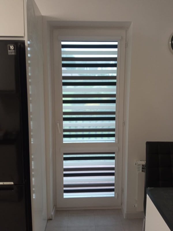 Okno z zamontowaną roletą dzień noc w kolorze szaro-białym. Widoczne poziome pasy rolety. Ujęcie z bliska, wnętrze mieszkania.