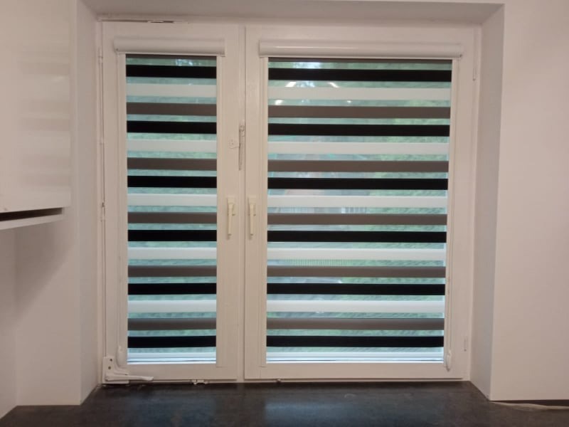 Dwuskrzydłowe okno z zamontowanymi roletami w dzień noc, z naprzemiennymi pasami w odcieniach szarości i bieli. Widoczne detale mechanizmu rolet.