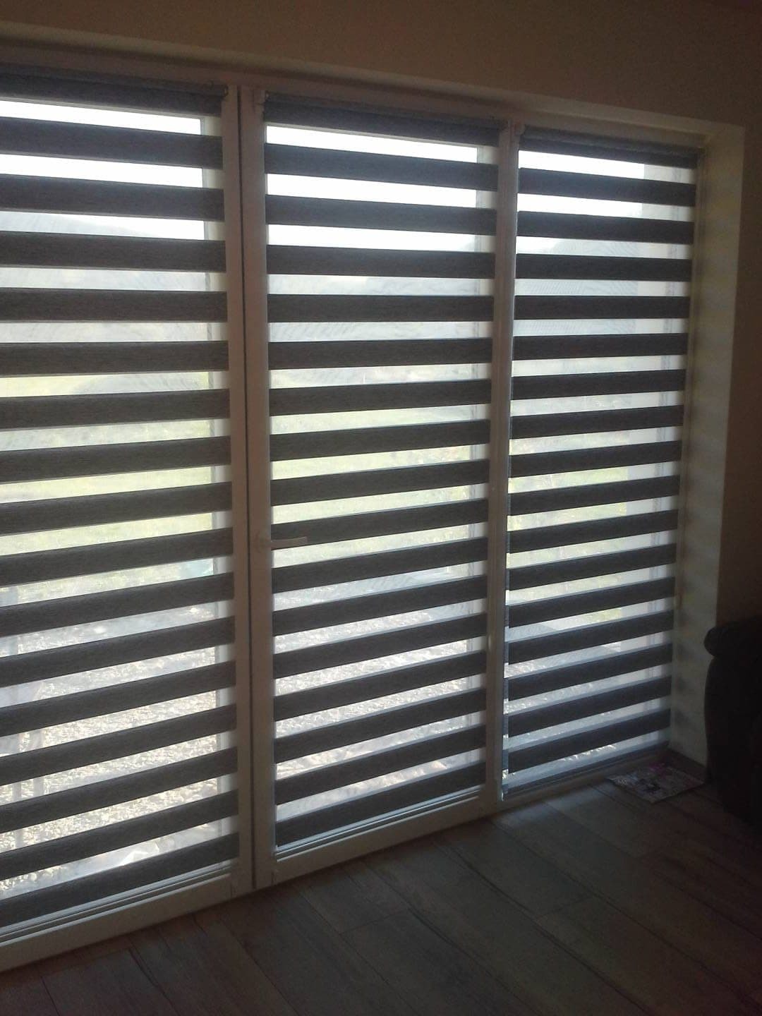 Trzy okna z zamontowanymi poziomymi przesłonami okiennymi w odcieniach szarości i bieli, tworzącymi efekt zebry, z widoczną strukturą materiału i delikatnym światłem.