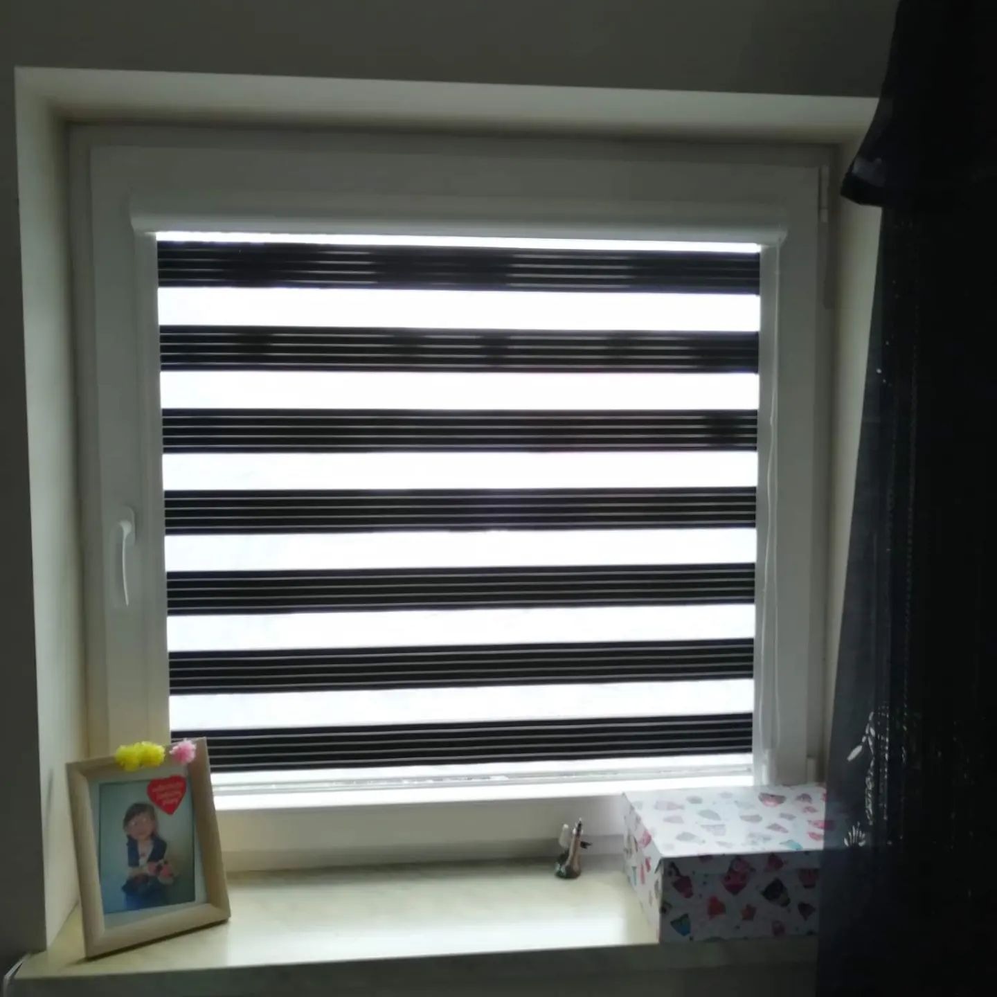 Okno z zamontowaną roletą zebra w biało-czarne pasy. Na parapecie ramka ze zdjęciem i pudełko w serduszka. Widok od wewnątrz, w tle ściana i zasłona.
