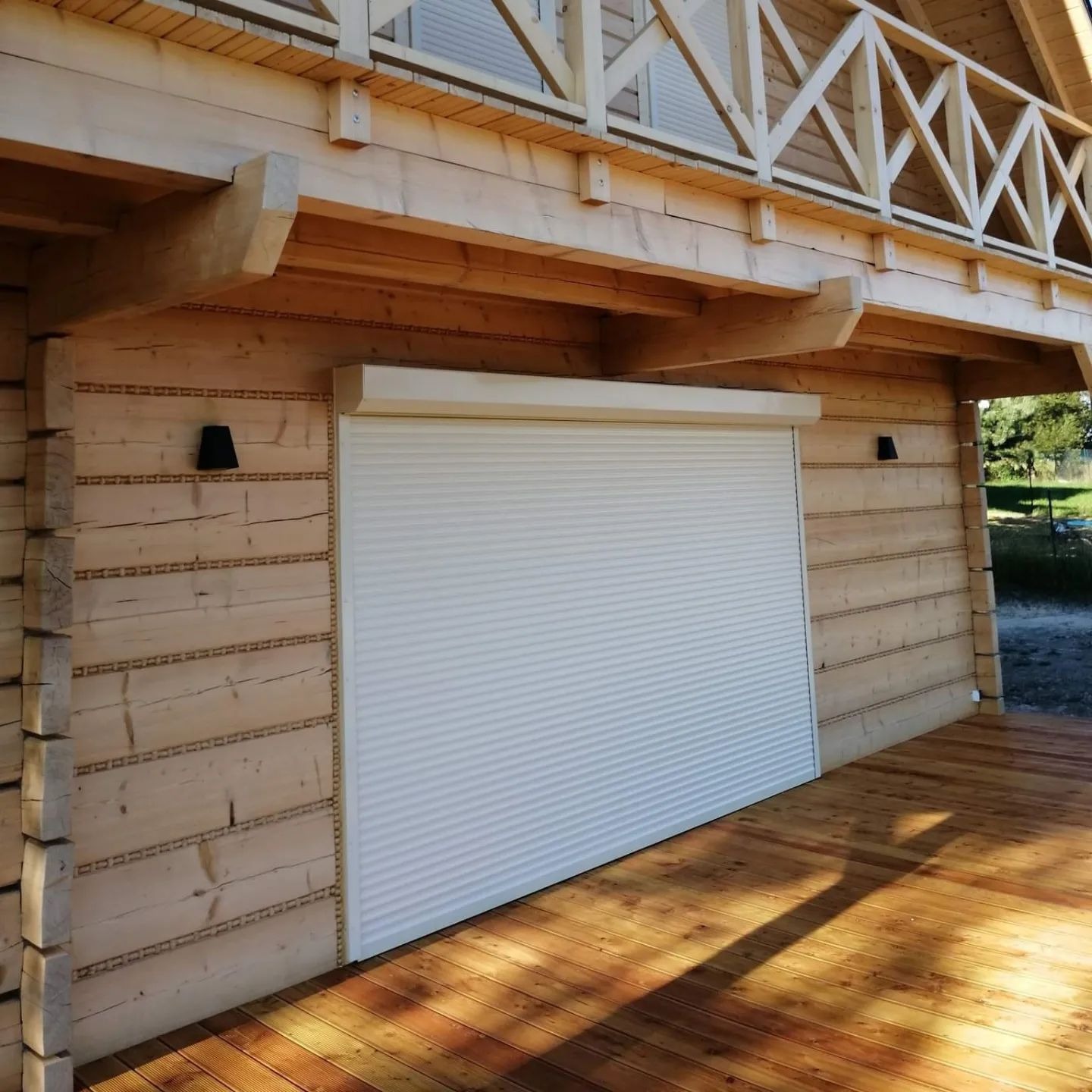 Zamknięta, biała roleta zewnętrzna na drewnianym budynku z balkonem. Widoczne drewniane deski i elementy konstrukcyjne. Ciepłe oświetlenie słoneczne.