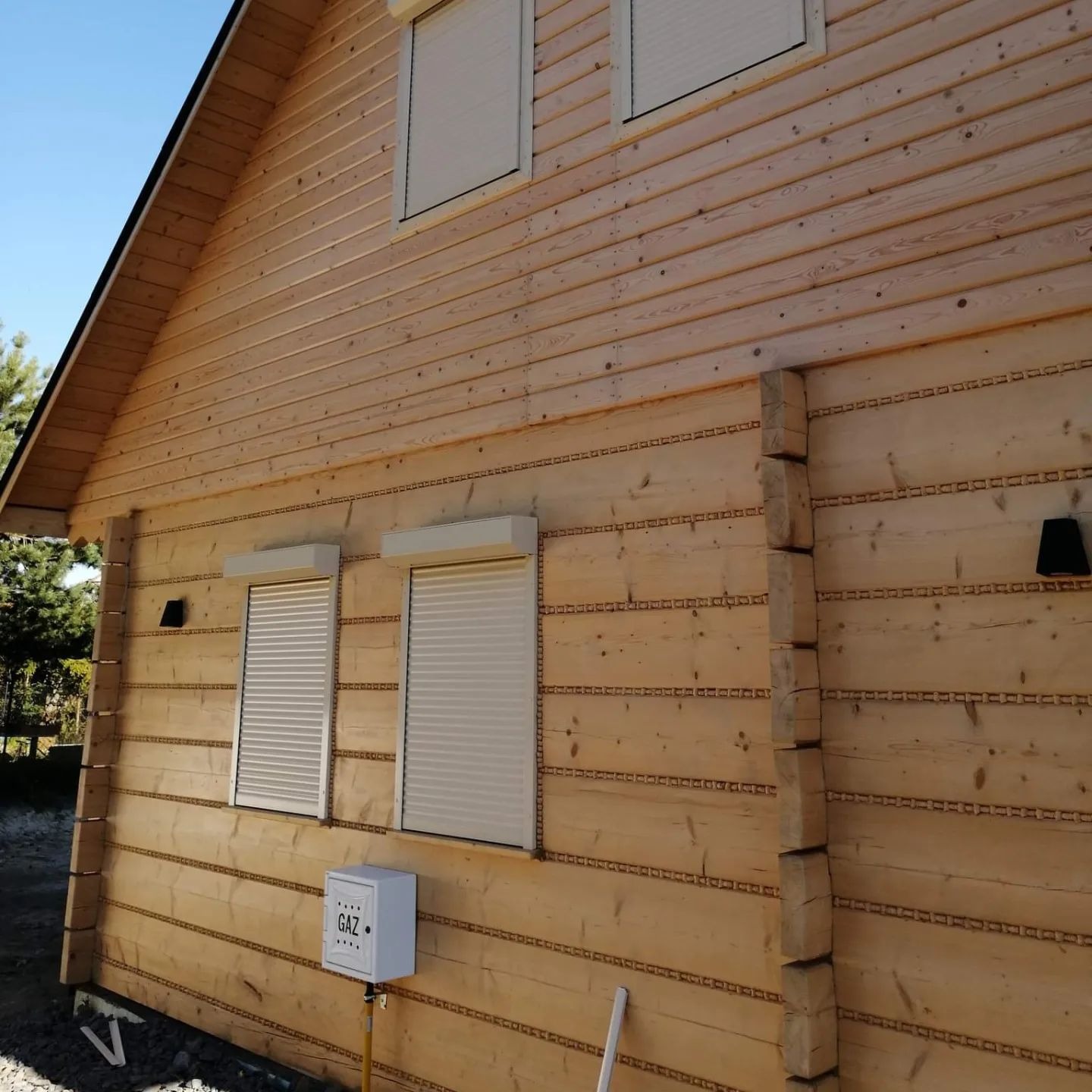 Drewniany dom z jasnymi, zamkniętymi osłonami okiennymi. Widoczne detale drewnianej elewacji i skrzynka gazowa przy ścianie. Słoneczny dzień.