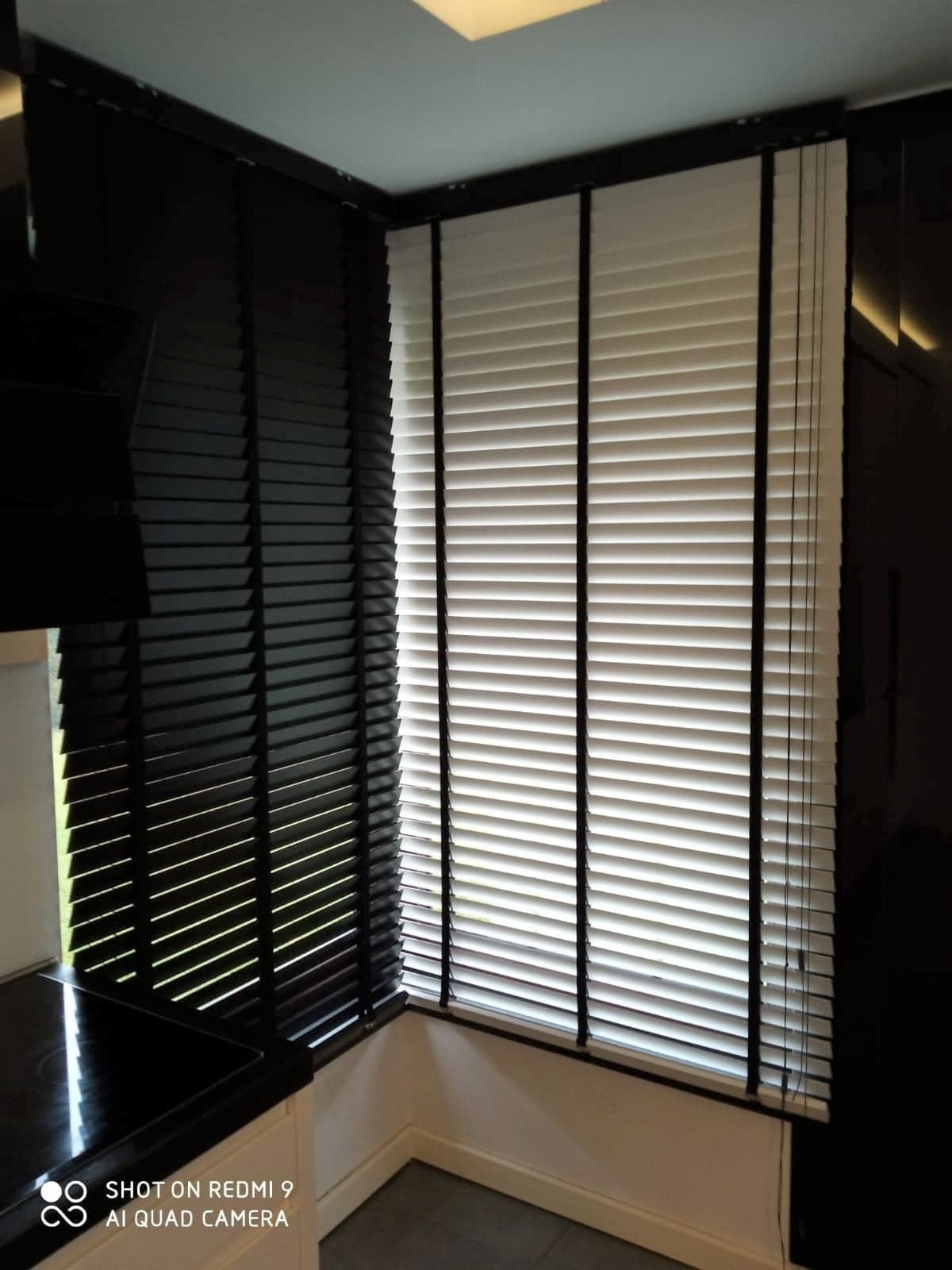 Zamknięte, poziome, drewniane przesłony okienne, białe z czarnymi taśmami, zamontowane w rogu okna w kuchni z czarnymi elementami wystroju.