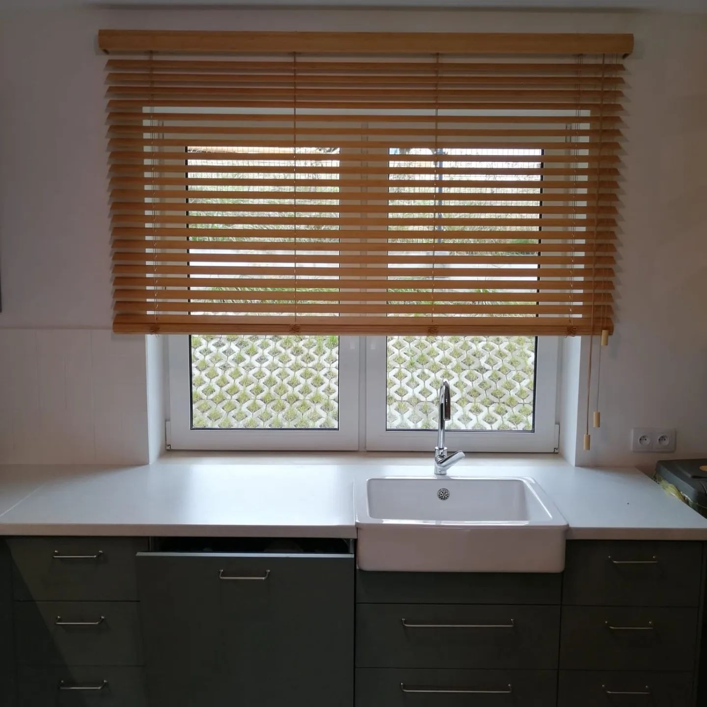 Kuchnia z drewnianymi żaluzjami poziomymi w oknie. Widok na zewnątrz przez żaluzje. Umywalka ceramiczna pod oknem. Szare szafki kuchenne.