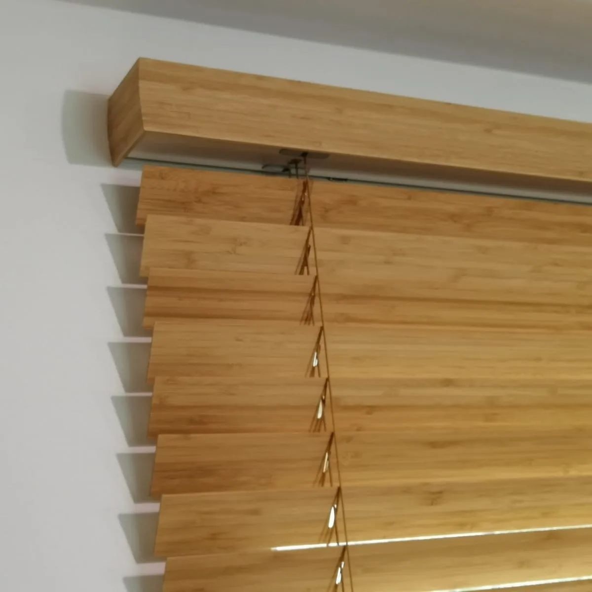 Jasne, drewniane żaluzje poziome zamontowane wewnątrz pomieszczenia. Widoczne lamele i mechanizm sterujący. Minimalistyczny design.