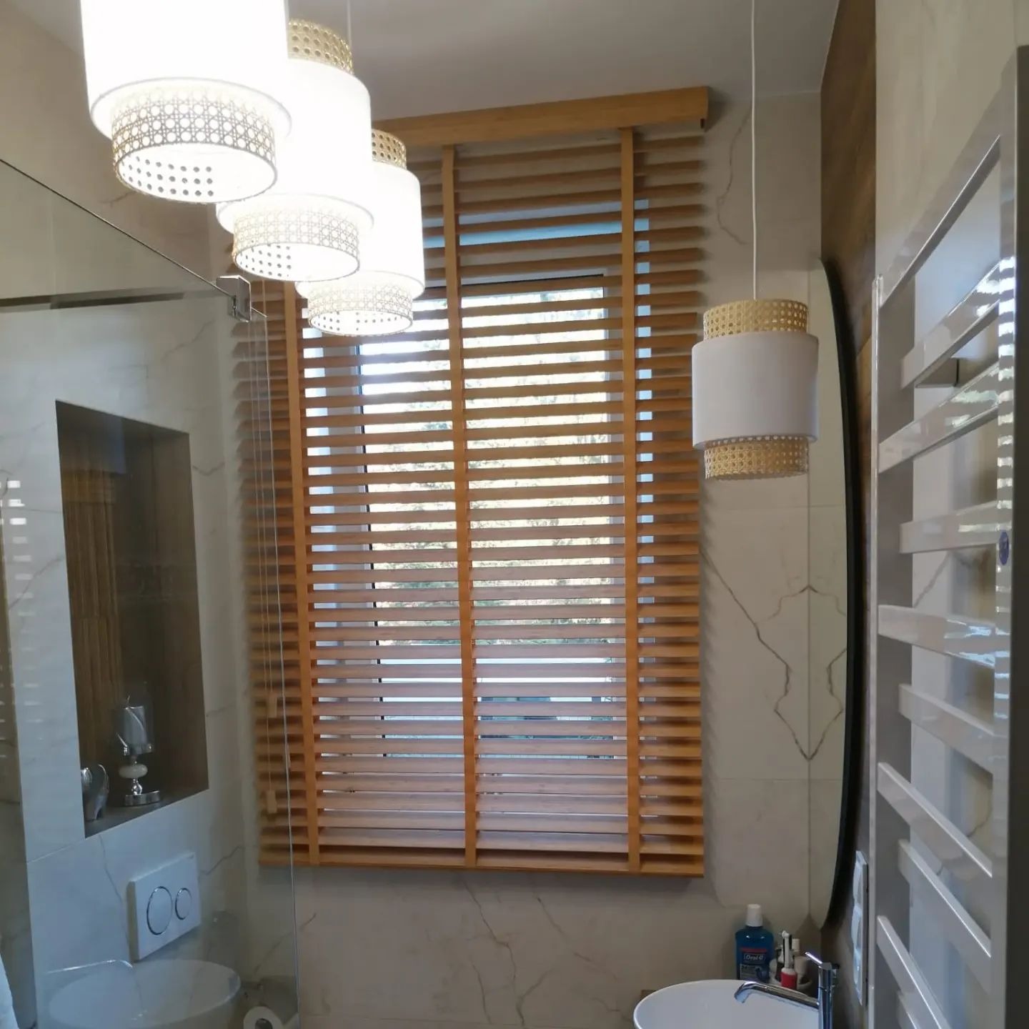 Stylowa łazienka z drewnianymi żaluzjami poziomymi, przez które wpada naturalne światło. Widoczne lampy wiszące i marmurowe płytki.