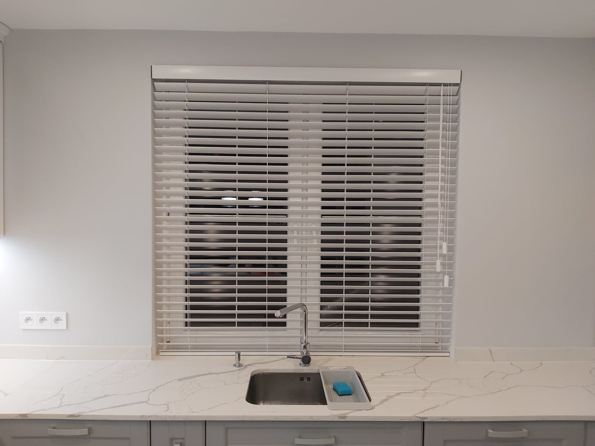 Białe żaluzje poziome zamontowane w oknie nad zlewem w jasnej kuchni. Widoczne gniazdka elektryczne i marmurowy blat.