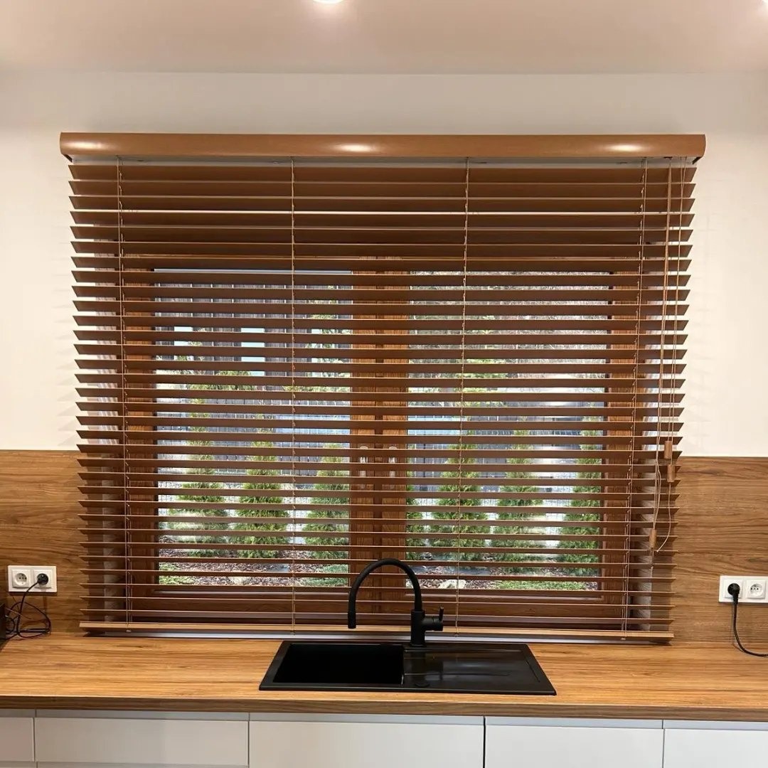Poziome drewniane żaluzje w oknie nad zlewem, w ciepłym odcieniu brązu, z widokiem na zieleń ogrodu, oświetlone punktowo od góry.